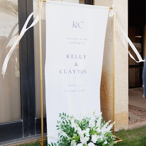 K&ouml;nnte beinhalten: Ein wei&szlig;es Hochzeitsbanner mit goldenem Metallrahmen, 91x203 cm, zeigt die Namen Kelly & Clayton. Das Banner ist mit wei&szlig;en Blumen und Gr&uuml;n am Fu&szlig; verziert und tr&auml;gt den Text "welcome to the wedding of".