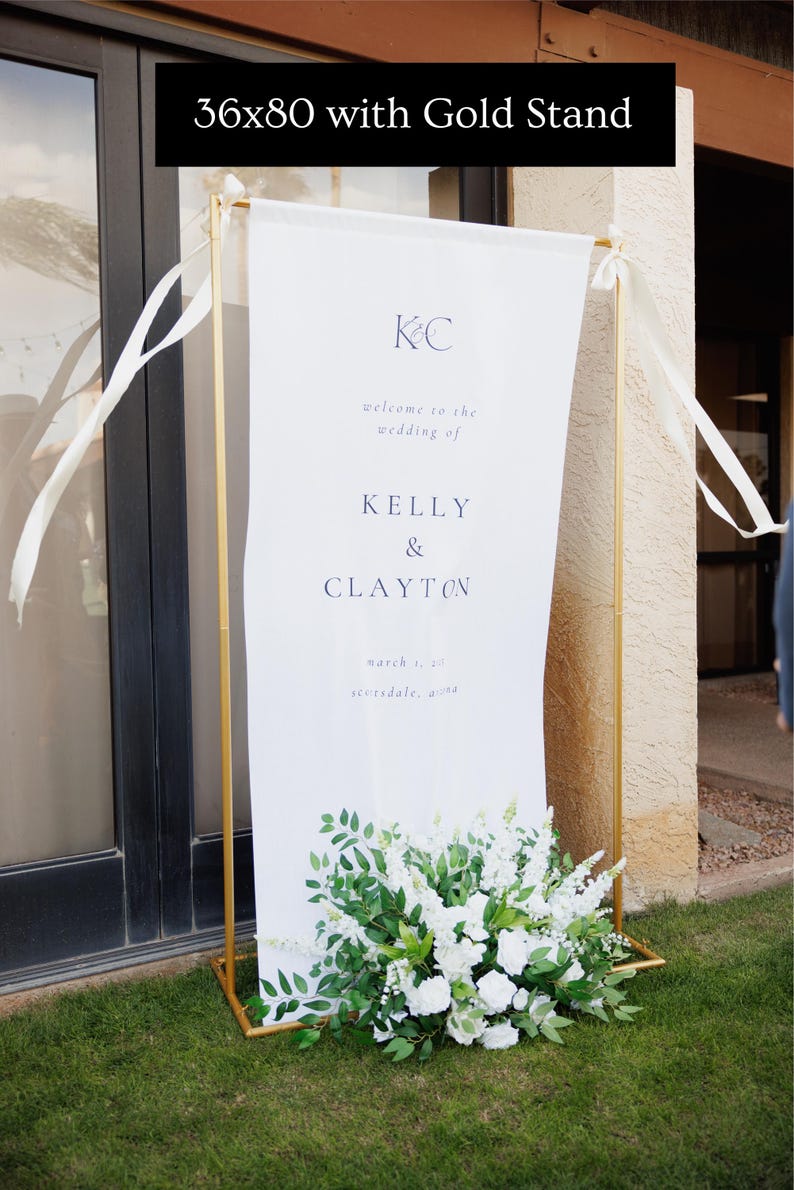 K&ouml;nnte beinhalten: Ein wei&szlig;es Hochzeitsbanner mit goldenem Metallrahmen, 91x203 cm, zeigt die Namen Kelly & Clayton. Das Banner ist mit wei&szlig;en Blumen und Gr&uuml;n am Fu&szlig; verziert und tr&auml;gt den Text "welcome to the wedding of".