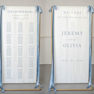 May include: Two white fabric wedding signs with blue text and light blue ribbon accents, displayed on gold metal stands. One sign reads "OUR FAVORITE PEOPLE" and lists guest names by table, while the other reads "WELCOME TO THE WEDDING OF JEREMY & OLIVIA".