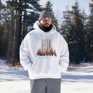 Puede incluir: Una sudadera con capucha blanca con un estampado de silueta de bosque marrón. El modelo lleva una boina gris y está de pie en un bosque nevado.