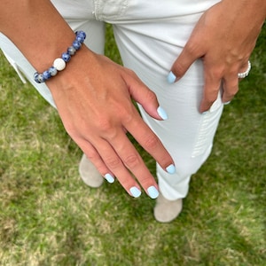 May include: A blue and white beaded bracelet with a white lava bead. The bracelet is on a person's wrist. The person is wearing white pants and has light blue nail polish.