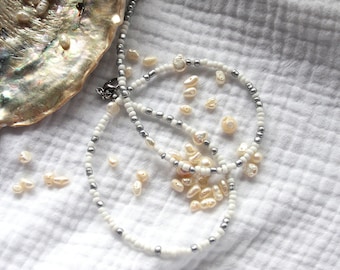May include: Two white and silver beaded necklaces with a pearl-like finish, scattered on a white fabric background with loose pearls.