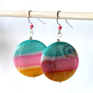 Könnte beinhalten: Ein Paar runde, farbenfrohe Ohrringe mit einem türkisfarbenen, weißen, rosa und gelben Streifenmuster. Die Ohrringe sind aus einem glatten, glänzenden Material gefertigt und hängen an silbernen Haken.
