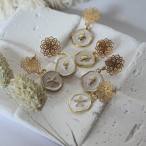 Può includere: Orecchini dorati a tema costiero. Gli orecchini presentano un delicato design a filigrana e ciondoli con motivi marini, tra cui stelle marine, conchiglie e tartarughe. I ciondoli hanno uno sfondo bianco e un bordo dorato.