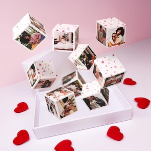 Puede incluir: Cubos de fotos blancos flotantes con imágenes de personas, algunas con vestidos de novia, otras abrazándose. Los cubos tienen un patrón de pequeños corazones rosas. Decoraciones rojas en forma de corazón esparcidas.
