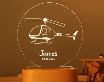 May include: A personalized night light featuring a helicopter design. The light is etched into a clear acrylic circle, with the name "James" and a date below. The base is made of wood. A wooden toy airplane is in the background.