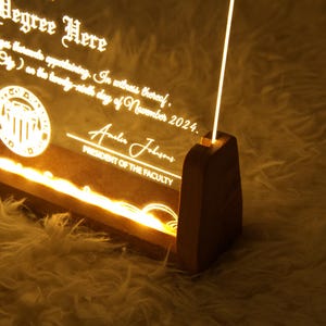 K&ouml;nnte beinhalten: Eine Holzplakette mit einem transparenten Acryleinsatz, beleuchtet mit warmwei&szlig;en LED-Lichtern. Die Plakette zeigt ein Siegel mit einem Geb&auml;udemotiv und dem Text "Degree Here". Der Text "Pr&auml;sident der Fakult&auml;t" und eine Unterschrift sind ebenfalls sichtbar.
