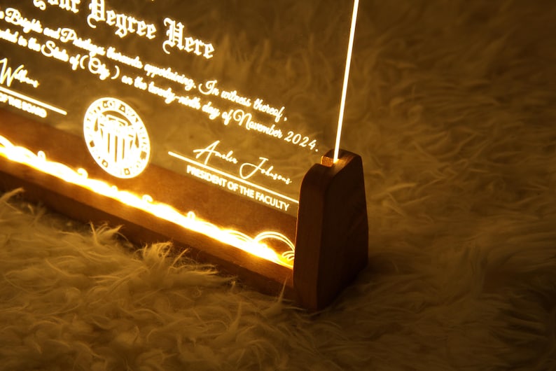 K&ouml;nnte beinhalten: Eine Holzplakette mit einem transparenten Acryleinsatz, beleuchtet mit warmwei&szlig;en LED-Lichtern. Die Plakette zeigt ein Siegel mit einem Geb&auml;udemotiv und dem Text "Degree Here". Der Text "Pr&auml;sident der Fakult&auml;t" und eine Unterschrift sind ebenfalls sichtbar.