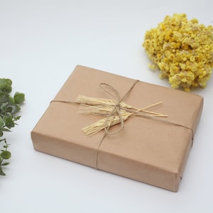 K&ouml;nnte beinhalten: Eine mit braunem Papier verpackte Geschenkbox, die mit Schnur gebunden und mit getrockneten Weizen&auml;hren verziert ist. Die Box steht auf einer wei&szlig;en Oberfl&auml;che mit gr&uuml;nen Eukalyptuszweigen und getrockneten gelben Blumen.