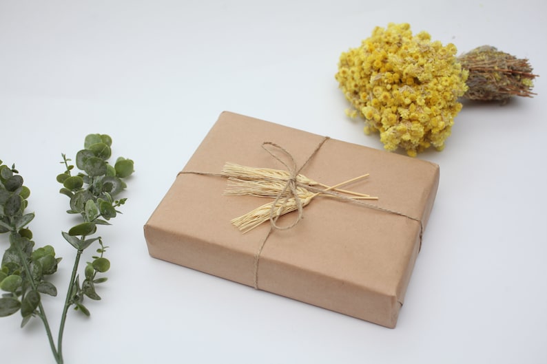K&ouml;nnte beinhalten: Eine mit braunem Papier verpackte Geschenkbox, die mit Schnur gebunden und mit getrockneten Weizen&auml;hren verziert ist. Die Box steht auf einer wei&szlig;en Oberfl&auml;che mit gr&uuml;nen Eukalyptuszweigen und getrockneten gelben Blumen.