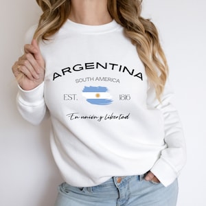 May include: White sweatshirt with a black graphic print that says "ARGENTINA" with the words "SOUTH AMERICA" and "EST. 1816" underneath. The graphic also includes a blue and white brushstroke design of the Argentinian flag with the text "En union y libertad" underneath.