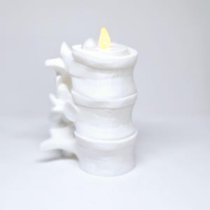 May include: A white ceramic candle holder shaped like a stack of bamboo with a small yellow flame on top.