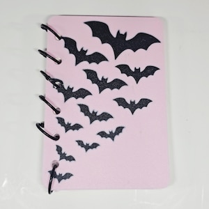 May include: A pink notebook with black spiral binding and a pattern of black bat silhouettes. The bats vary in size and are arranged in a descending diagonal pattern across the cover. The notebook is a rectangular shape.