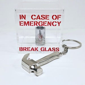 May include: A clear plastic box with the text "IN CASE OF EMERGENCY BREAK GLASS" printed in red. Inside the box is a silver socket wrench. A silver hammer keychain is attached to the box.