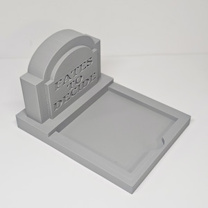 May include: A gray, 3D-printed tombstone-shaped desk organizer. The tombstone reads "Fates to Decide" in carved lettering. The organizer has a rectangular tray for holding items.