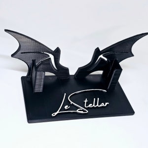 May include: Black 3D printed stand with two bat wings on either side. The stand has a black base with the signature "Le Stellar" written in white.