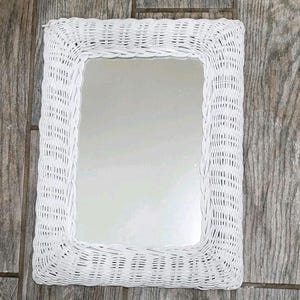 May include: A rectangular white wicker mirror. The mirror has a woven frame and a reflective surface. The frame is made of tightly woven white wicker, creating a textured border around the glass. The mirror is designed for wall mounting.