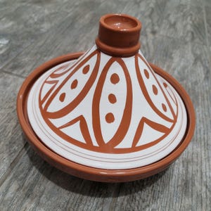 May include: A white ceramic tagine lid with a brown geometric pattern. The lid is round and has a small opening in the center.