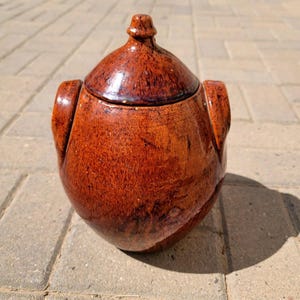 May include: A handcrafted, brown ceramic jar with a lid. The jar has a rounded body with two side handles and a decorative knob on the lid. The jar's surface has a glossy finish and a rich, earthy tone, suggesting a rustic or vintage aesthetic.