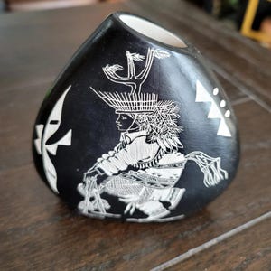 May include: Black ceramic vase with a white interior and a detailed carved design. The design features a figure in traditional attire, with intricate patterns and geometric shapes. The vase has a unique, rounded shape and a small opening at the top.