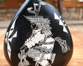 May include: A smooth, black, teardrop-shaped stone featuring a detailed white etched design of a figure in traditional regalia with a feathered headdress, holding a bundle of arrows, with geometric patterns on the sides of the stone.