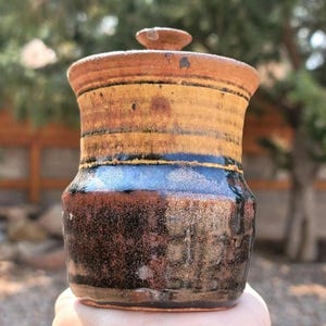 May include: A handmade ceramic jar with a lid. The jar features a textured surface with a color gradient of brown, yellow, and black. The jar is held in a hand.