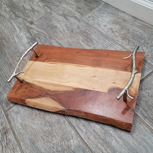 May include: A wooden serving tray with a natural wood grain finish and two silver metal handles shaped like branches. The tray is rectangular with rounded edges.