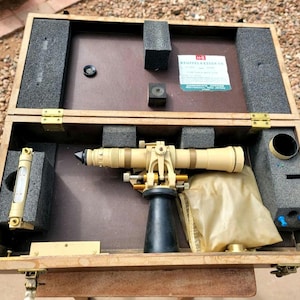 May include: A vintage Keuffel & Esser surveying instrument housed in a wooden carrying case with foam inserts. The set includes a tan telescope with a black base, a spirit level, and a translucent protective cover, all secured within the box.