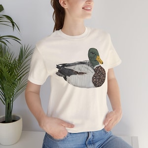 Può includere: Una t-shirt a maniche corte color crema con una grafica centrale raffigurante un germano reale. L'anatra è disegnata in stile patchwork, incorporando vari motivi di tessuto tra cui tartan verde, fiori marroni e sezioni a righe grigio chiaro.
