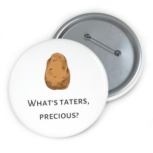 May include: White button pin with a brown potato illustration and the text "WHAT'S TATERS, PRECIOUS?". The pin has a metal backing and a safety pin closure. The design is a humorous reference to a popular fantasy novel.