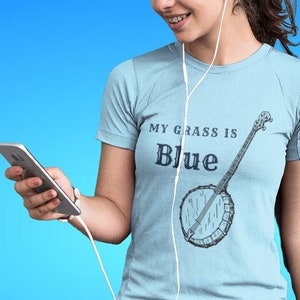 May include: A woman wearing a light blue t-shirt with the text "My grass is blue" and a banjo illustration. She is wearing black leggings and white headphones.