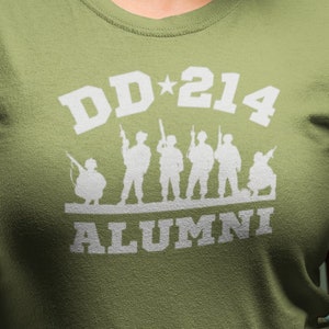 May include: A woman wearing a green t-shirt with the text "DD*214 ALUMNI" and a white silhouette of soldiers. She is holding a silver dog tag necklace.