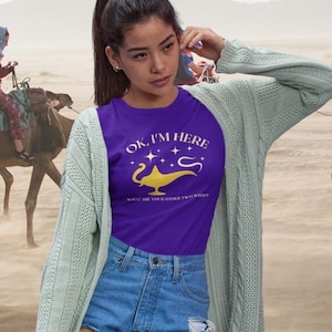Op de afbeelding: Een vrouw draagt een paars T-shirt met de tekst "OK, I'M HERE" en een gouden djinn-lamp afbeelding. Ze draagt een lichtblauwe cardigan en een spijkerbroek. De achtergrond is een woestijnscène met kamelen.