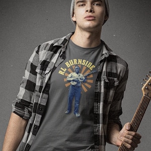 Puede incluir: Una persona con una camiseta gris con el texto "RL Burnside" y un gráfico de un músico. La persona sostiene una guitarra eléctrica roja. Viste una camisa a cuadros, vaqueros y botas.
