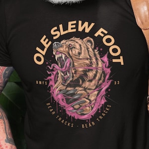 May include: Black t-shirt with a graphic of a bear with the text "Ole Slew Foot" in a circle around it. The text "BMFS" and "33" are also included in the circle. The text "Bear Tracks" is written twice below the circle.