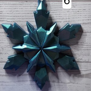 May include: A teal-colored, geometric snowflake ornament with a shimmering finish, hanging by a white string. The number 6 is visible in the background. The ornament is made of a hard material and has sharp edges.