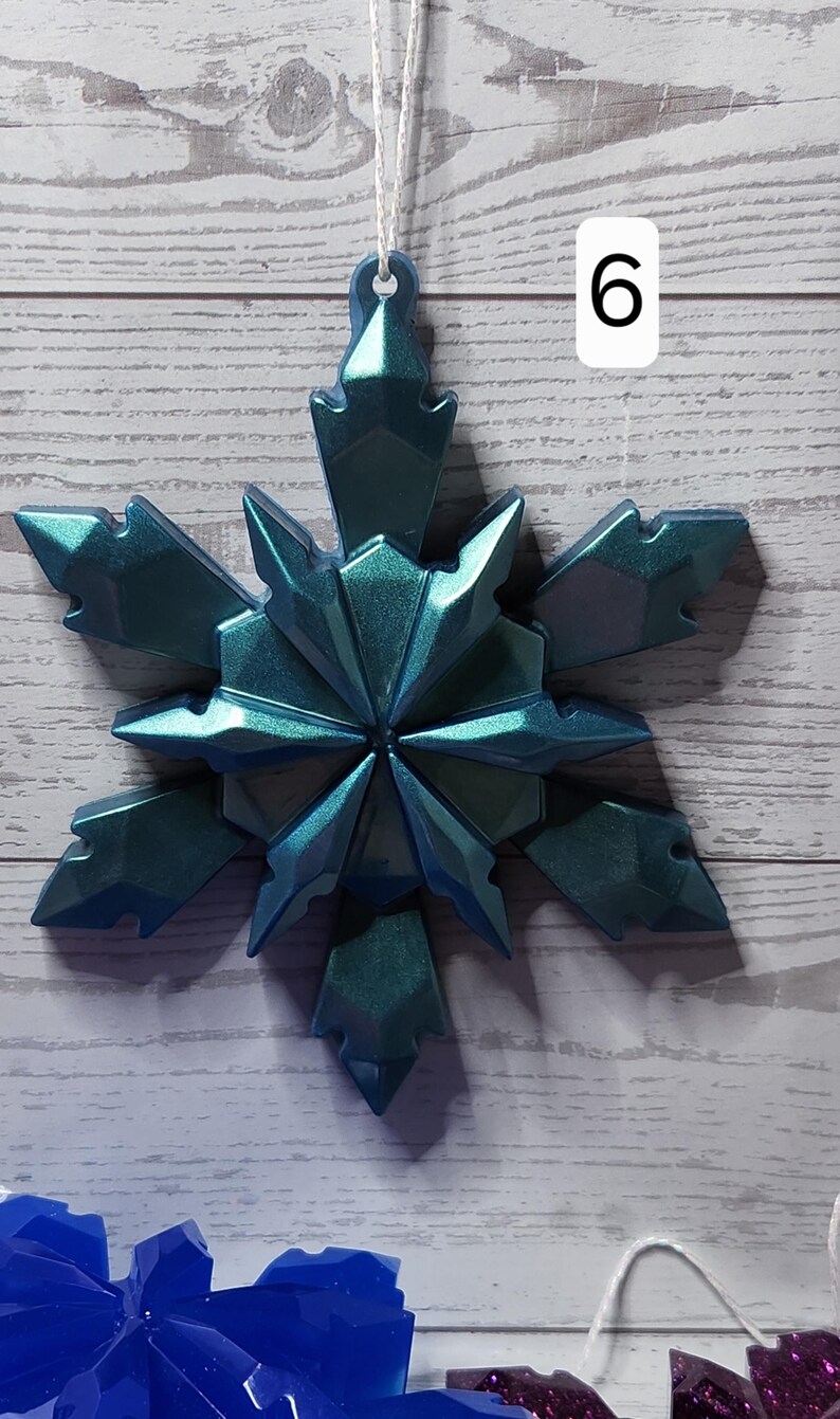 May include: A teal-colored, geometric snowflake ornament with a shimmering finish, hanging by a white string. The number 6 is visible in the background. The ornament is made of a hard material and has sharp edges.
