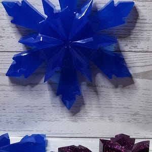 May include: A vibrant blue snowflake ornament with a faceted design, hanging from a white string. The number "2" is visible in the background. Other snowflake ornaments in shades of blue and purple are also visible.