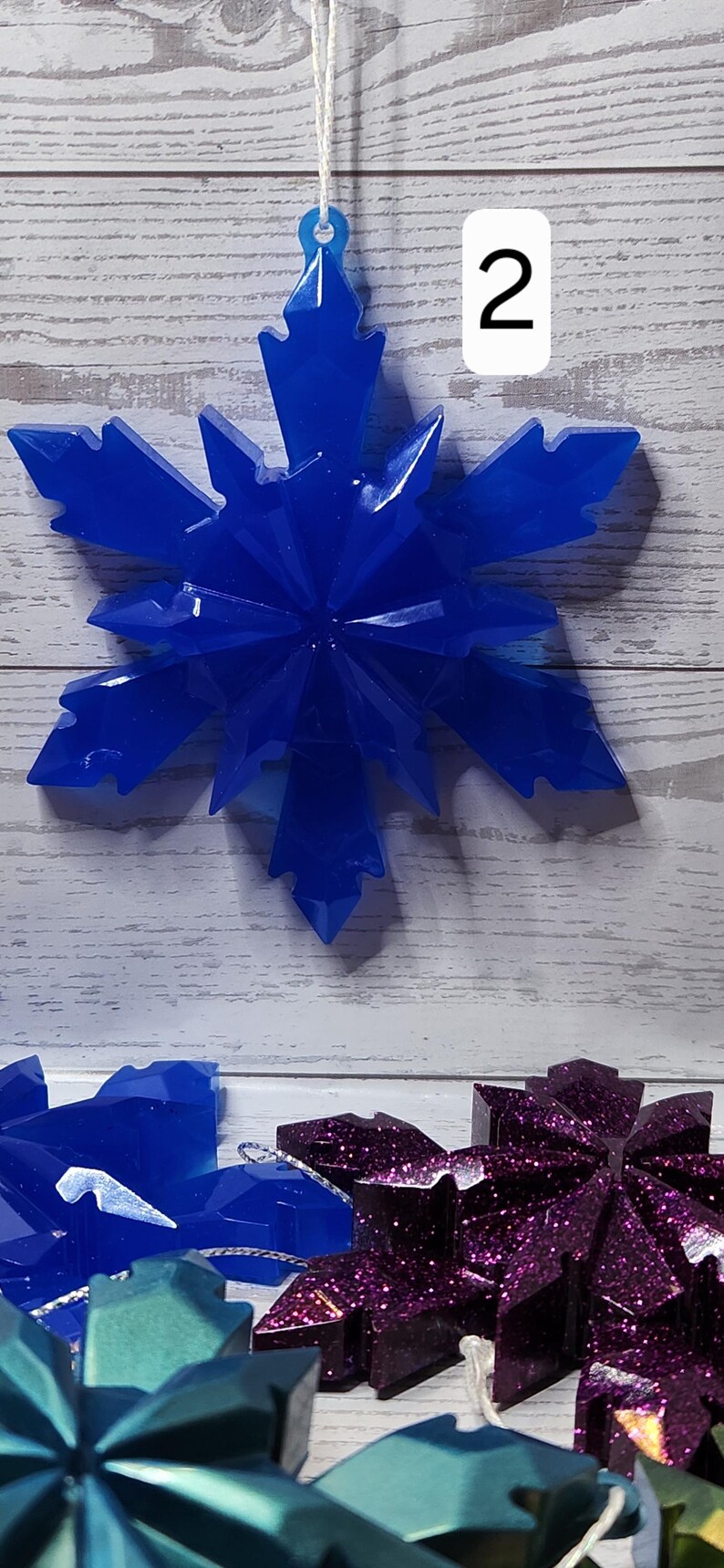 May include: A vibrant blue snowflake ornament with a faceted design, hanging from a white string. The number "2" is visible in the background. Other snowflake ornaments in shades of blue and purple are also visible.