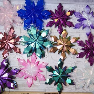 May include: A collection of decorative snowflake ornaments in various colors, including pink, blue, purple, and green. Each snowflake has a faceted design and is strung with a white cord for hanging. The ornaments are arranged on a light-colored wooden surface.