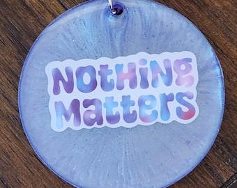 May include: A purple and white resin keychain with a paw print charm and a purple tassel. The keychain features a white sticker with the text "Nothing Matters" in a colorful tie-dye font.