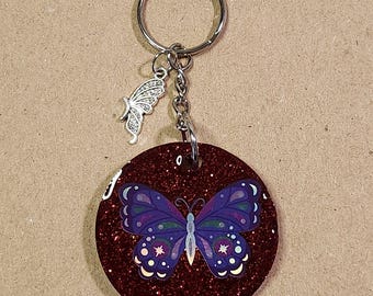 May include: A round, glittery burgundy keychain with a purple butterfly design. The butterfly has blue and white accents. A small silver butterfly charm hangs from the keychain ring.