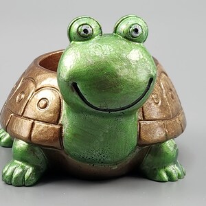 May include: A whimsical turtle-shaped planter with a smiling face. The turtle has a green head and feet, a brown and gold shell, and a circular opening for planting. The planter is made of a durable material.