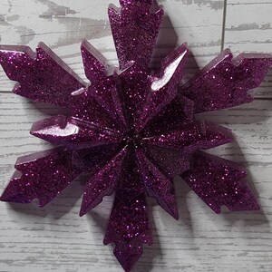 May include: A glittery purple snowflake ornament with a white string for hanging. The snowflake has a multi-pointed design and is covered in sparkling glitter. The number 8 is in a white rectangle in the background.