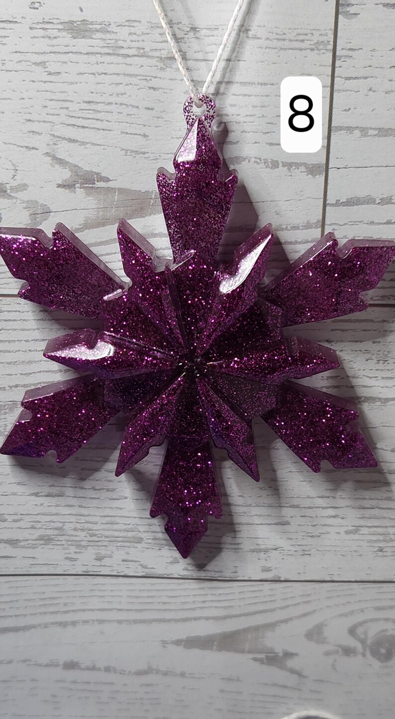 May include: A glittery purple snowflake ornament with a white string for hanging. The snowflake has a multi-pointed design and is covered in sparkling glitter. The number 8 is in a white rectangle in the background.