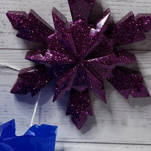 May include: A glittery purple snowflake ornament with a white string for hanging. The number "3" is on a white square in the background. The ornament is on a light wood background.