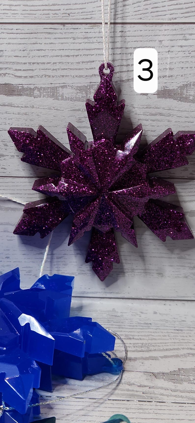 May include: A glittery purple snowflake ornament with a white string for hanging. The number "3" is on a white square in the background. The ornament is on a light wood background.