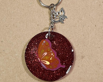 May include: A red glitter keychain with a silver butterfly charm. The keychain features a red glitter background with a colorful butterfly design.