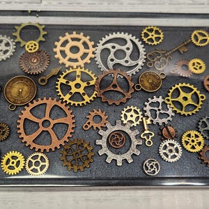 May include: A collection of various sized gears and keys in shades of gold, silver, and copper, set in a clear resin tray. The gears have intricate designs, creating a steampunk aesthetic.