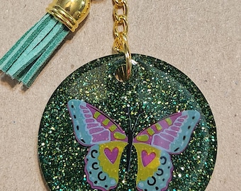 May include: A green glitter keychain with a gold chain and a green tassel. A pink and yellow butterfly with hearts is embedded in the glitter.